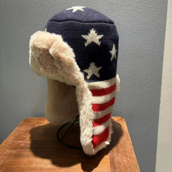 American flag patterned hat with fur trim on a wooden block against a gray wall.