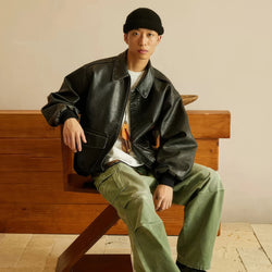 Person wearing a black leather jacket and green pants sitting on a wooden bench.